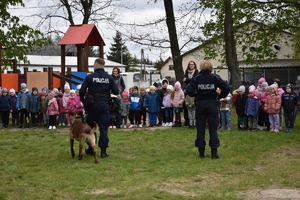 policjant przewodnik psa z policjantką stoją na placu zabaw, w tle dzieci